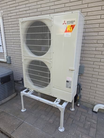 A Mitsubishi Zuba heat pump installed on a stand beside a brick house